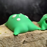 Back view of a green 3D printed blob frog with white spots, featuring its rounded, squishy-looking body and tiny leg nubs resting on a rock.