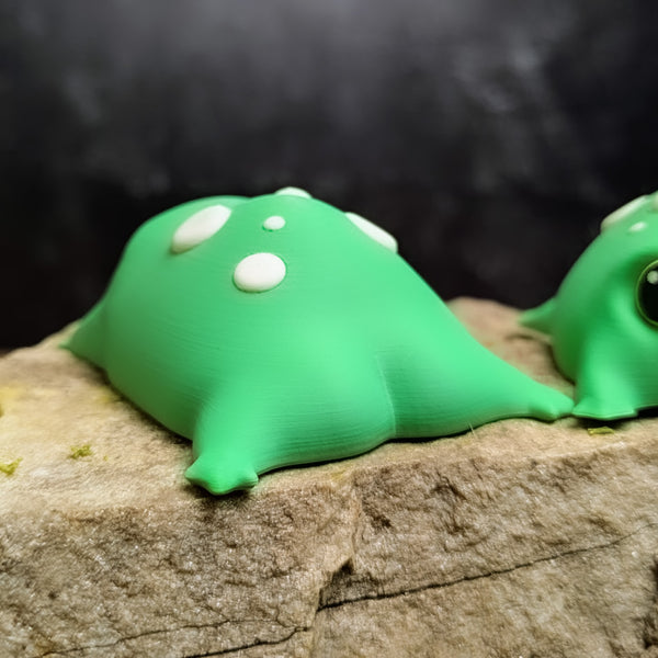 Back view of a green 3D printed blob frog with white spots, featuring its rounded, squishy-looking body and tiny leg nubs resting on a rock.