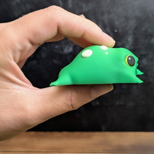 The blob frog held gently between fingers, viewed from the side, showcasing its soft form and simple spot details.