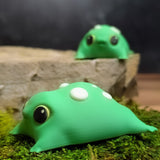 Side profile of the blob frog resting on moss, showing the rounded shape, white spots, and subtle foot nubs.