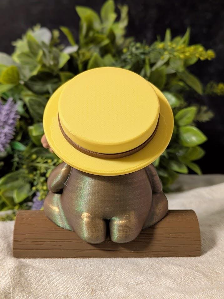 Back view of a whimsical frog figurine wearing a wide yellow sun hat and holding a tiny banjo.