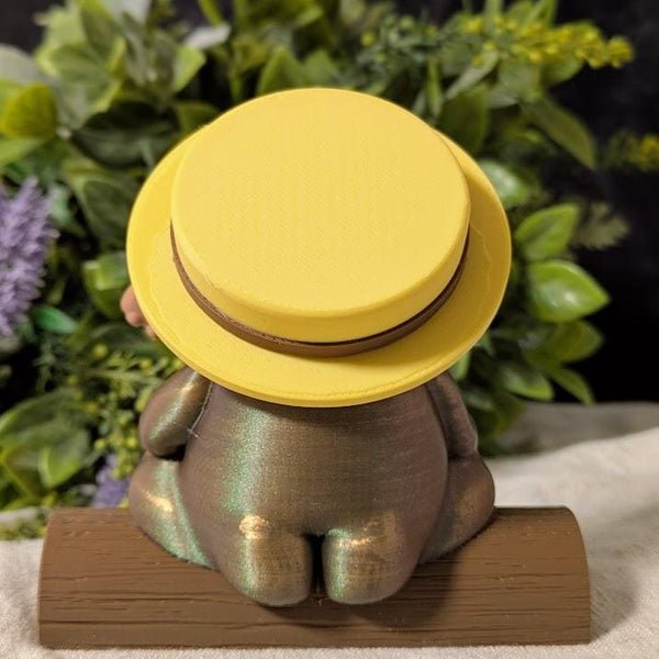 Back view of a whimsical frog figurine wearing a wide yellow sun hat and holding a tiny banjo.