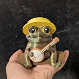 Frog figurine wearing a yellow hat, holding a tiny banjo while sitting on a human hand.