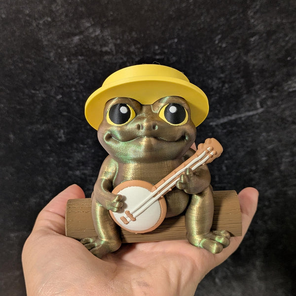 Frog figurine wearing a yellow hat, holding a tiny banjo while sitting on a human hand.