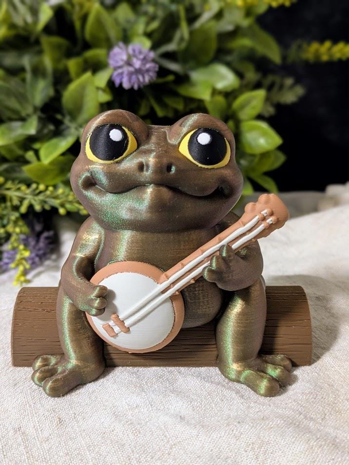 Frog figurine with a banjo and yellow hat sitting on a surface, surrounded by purple flowers, without a hat