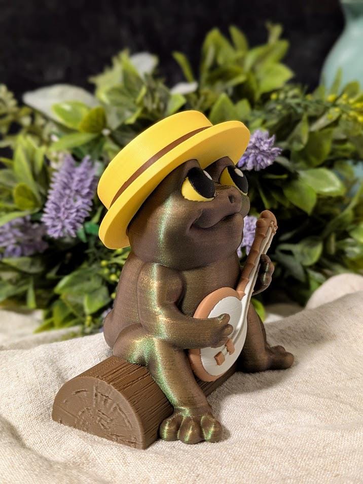 Side view of a frog figurine in a yellow hat, mid-banjo serenade, surrounded by leafy plants and flowers.