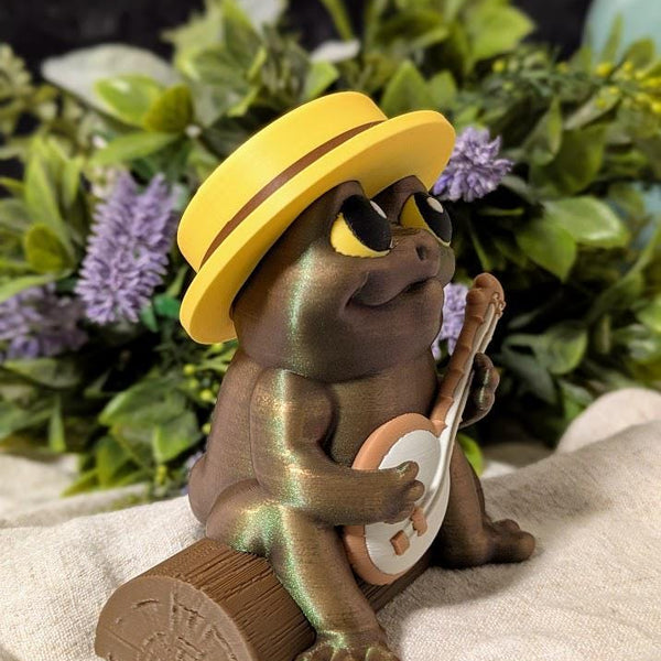 Side view of a frog figurine in a yellow hat, mid-banjo serenade, surrounded by leafy plants and flowers.