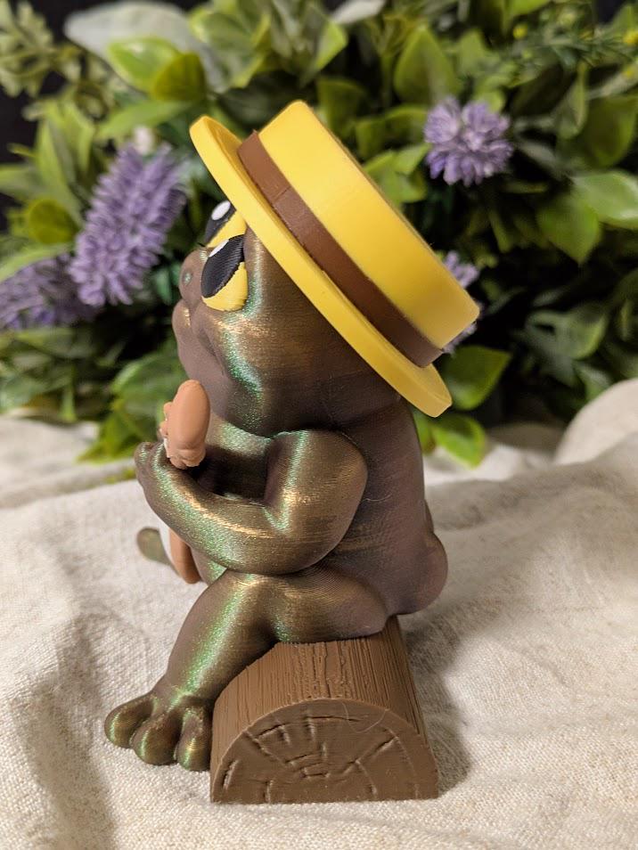 Side profile of frog figurine playing a banjo, wearing a yellow hat in a floral setting.