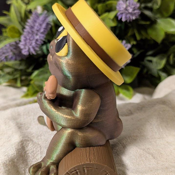 Side profile of frog figurine playing a banjo, wearing a yellow hat in a floral setting.