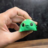 A hand holding the front of the blob frog, showing its expressive wide eyes and rounded body with soft sculpted detail.