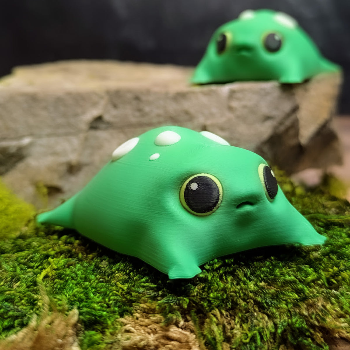 Front-facing view of a green blob frog figurine with big black eyes and white mushroom-like spots, sitting on green moss.