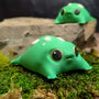 Front-facing view of a green blob frog figurine with big black eyes and white mushroom-like spots, sitting on green moss.