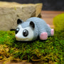 Front-facing blob possum figurine with wide cartoon eyes, gray fur texture, and pink feet and nose, resting on a mossy surface.