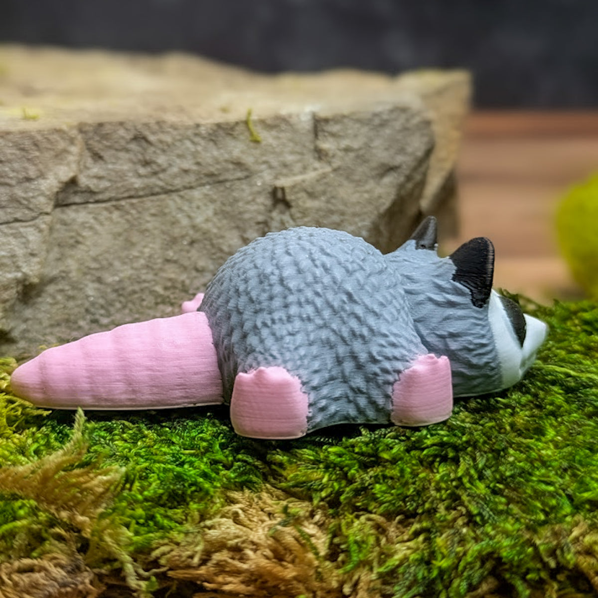 Side view of the blob possum on moss with its pink tail extended and head turned slightly forward.