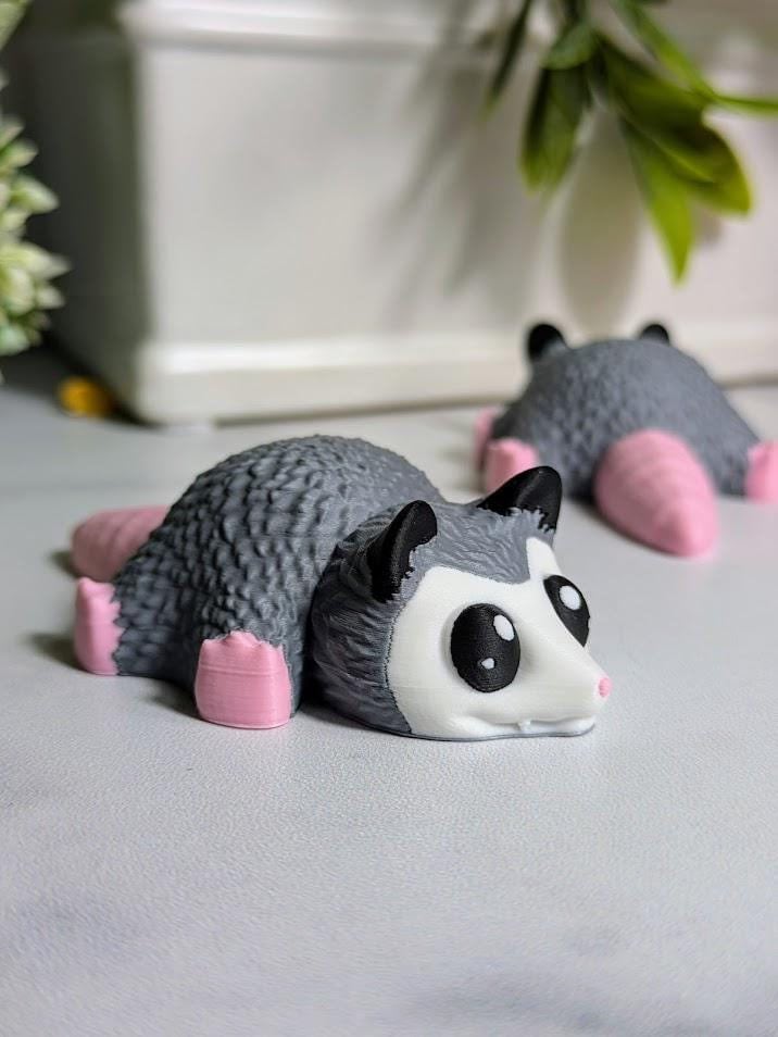A gray possum figurine with pink feet and tail lies on a white surface with a soft background.
