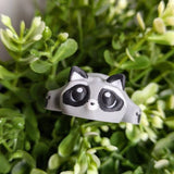 A blob raccoon peeks out from dense greenery, its big eyes staring upward with intense curiosity and a hint of chaos.