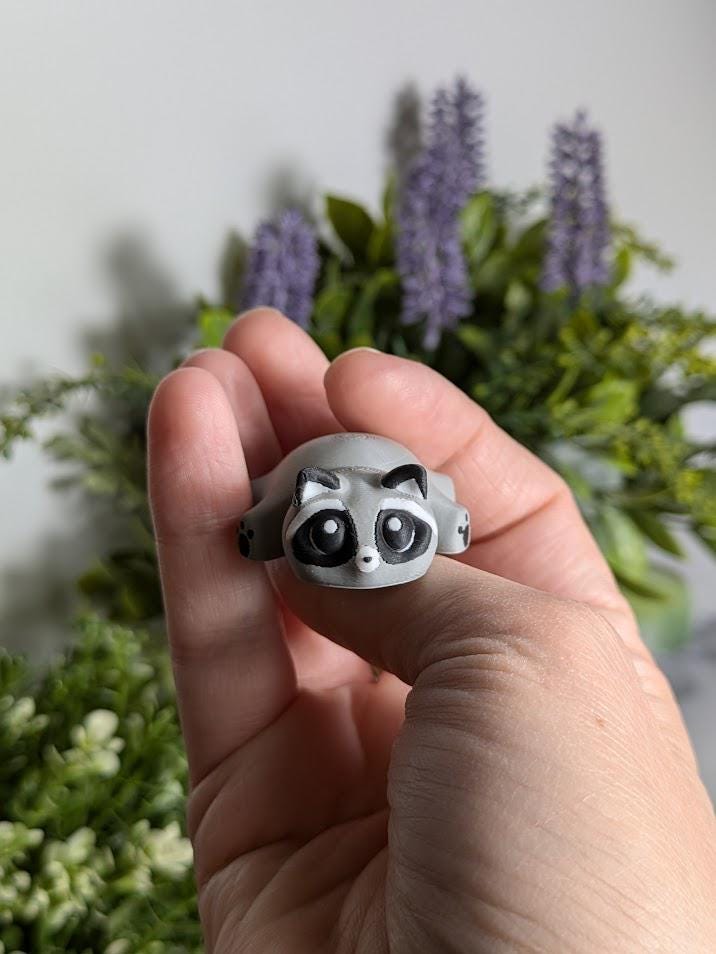 A front-facing view of the blob raccoon being held up by one hand, showing its rounded body, mask-like markings, and tiny snout.