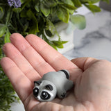 A soft, round blob raccoon figurine with wide eyes rests gently in a person's hand, surrounded by sprigs of lavender.