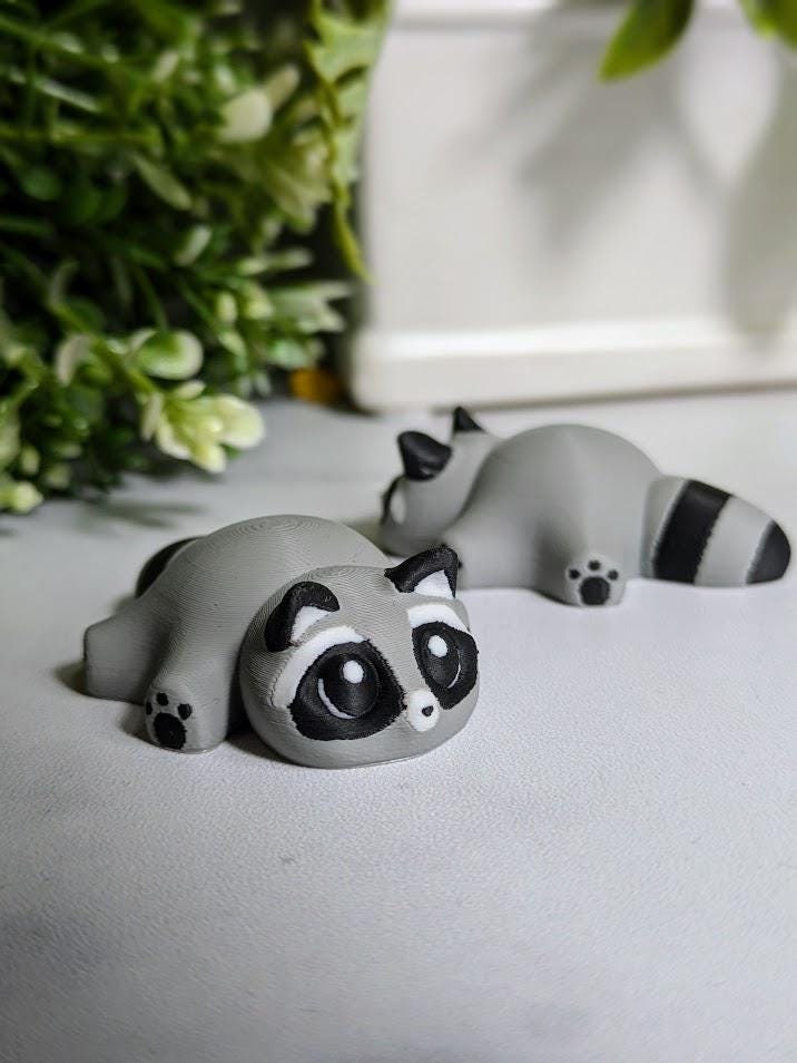 A side profile of a single blob raccoon shows off its stout body and striped tail as it sits peacefully on a white background.