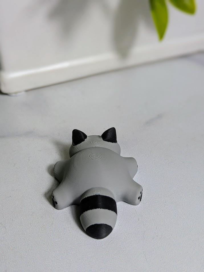A top-down view of the blob raccoon figurine lying on a white surface, showing its small ears, chubby shape, and striped tail.