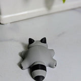 A top-down view of the blob raccoon figurine lying on a white surface, showing its small ears, chubby shape, and striped tail.
