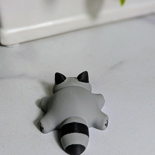 A top-down view of the blob raccoon figurine lying on a white surface, showing its small ears, chubby shape, and striped tail.