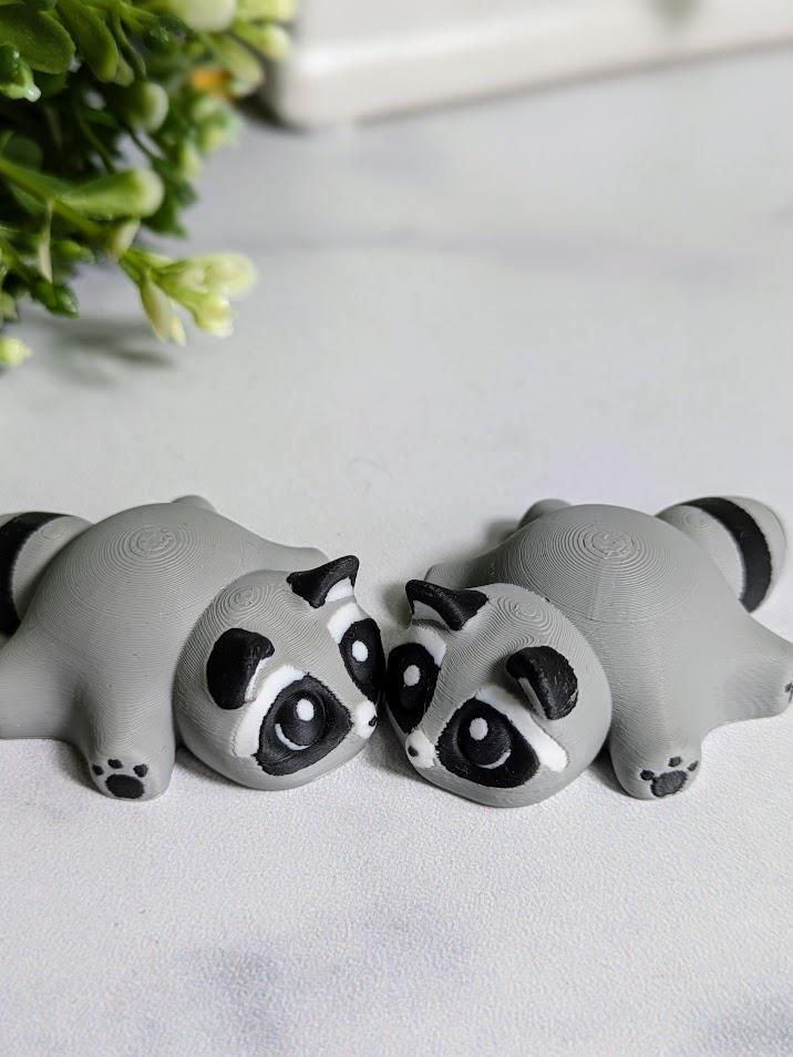 Two blob raccoon figurines sit side by side on a white surface, their wide-eyed expressions pointed slightly inward like they're sharing secrets.