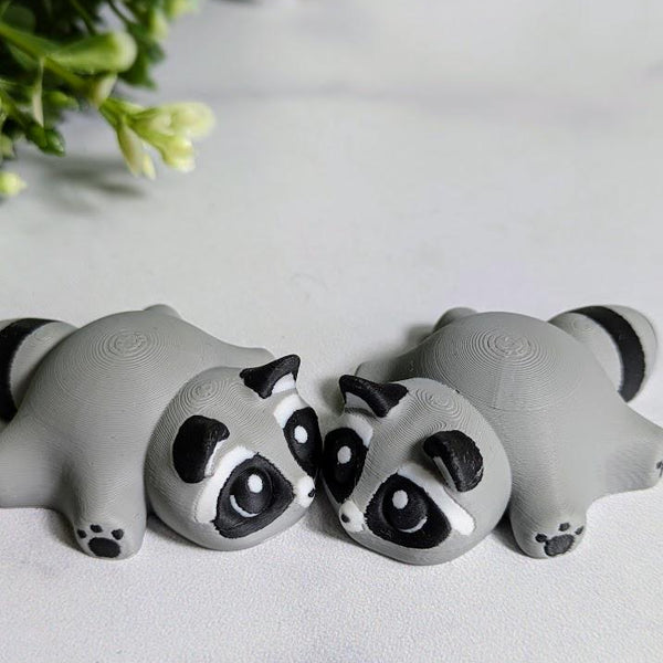 Two blob raccoon figurines sit side by side on a white surface, their wide-eyed expressions pointed slightly inward like they're sharing secrets.