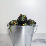Multiple frog figurines crammed together inside a silver metal bucket, evoking maximum judgment per ounce.