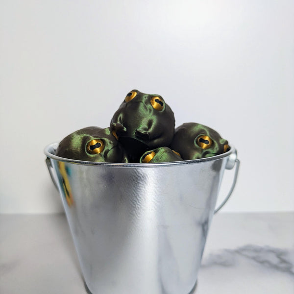 Multiple frog figurines crammed together inside a silver metal bucket, evoking maximum judgment per ounce.