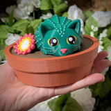 Green cactus cat figurine with flower, sitting in terracotta pot with wide-eyed expression.