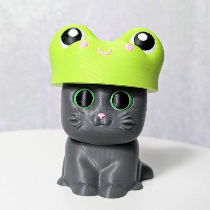 Gray cat figurine wearing a bright green frog hat with bulging cartoon eyes.