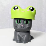 Gray cat figurine wearing a bright green frog hat with bulging cartoon eyes.