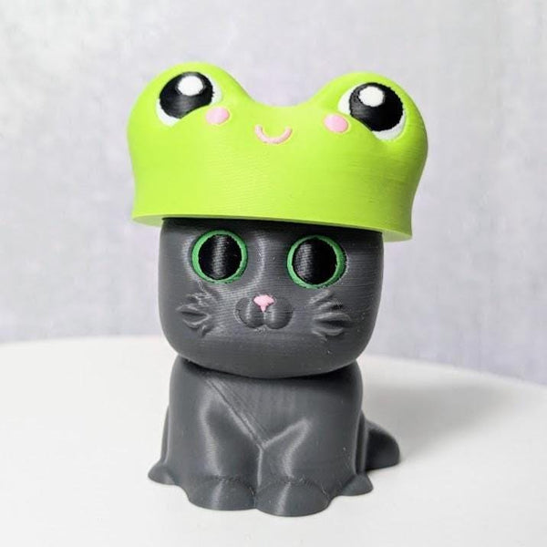 Gray cat figurine wearing a bright green frog hat with bulging cartoon eyes.