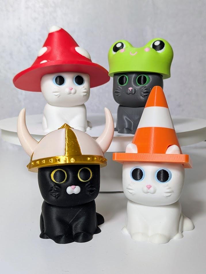Group of three cat figurines each wearing different hats: mushroom, frog, and traffic cone.