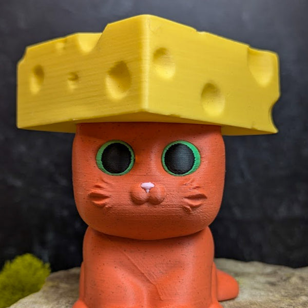 An orange cat figurine with large green eyes wearing a blocky, wedge-shaped yellow cheese hat.