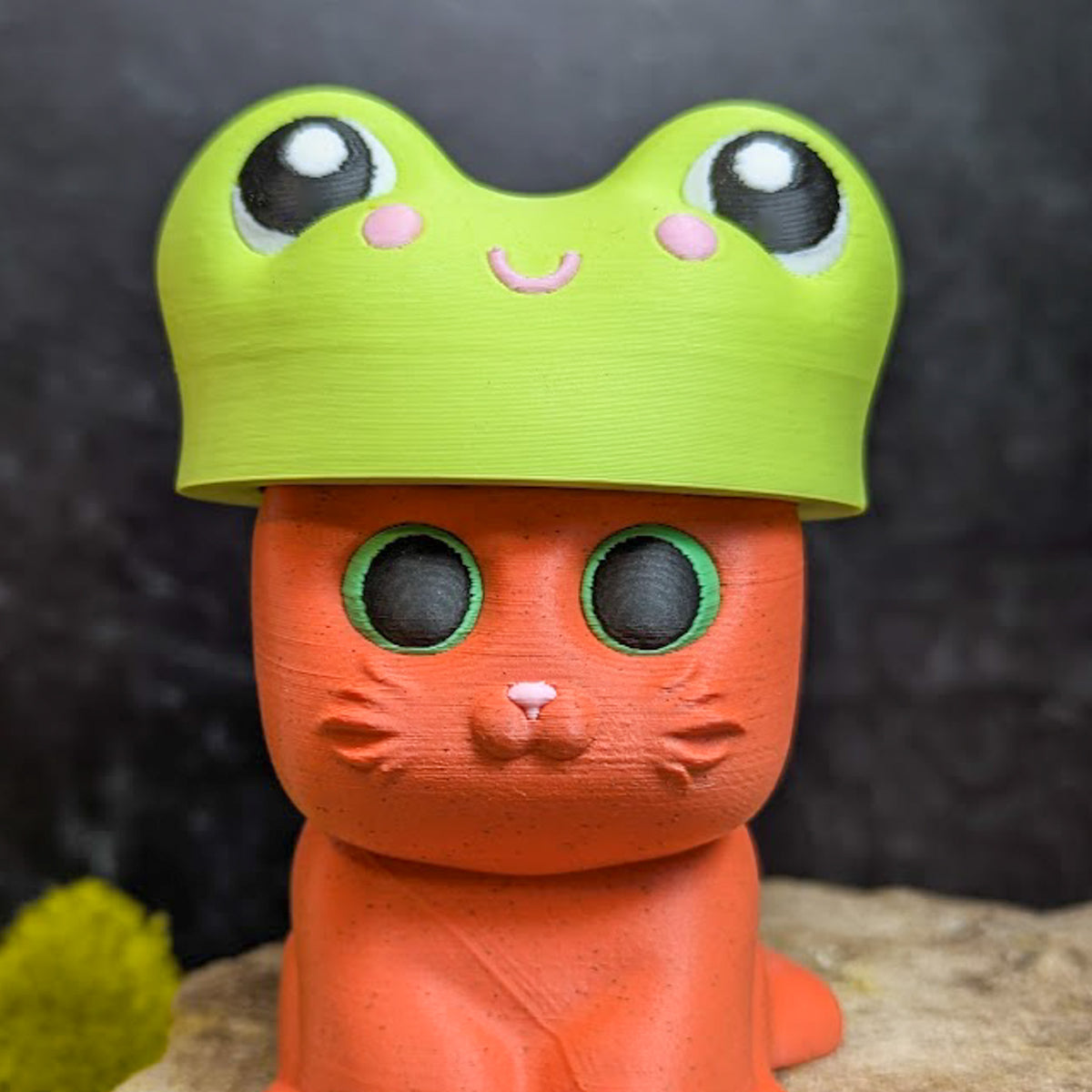 An orange cat figurine with green eyes wearing a bright green frog hat featuring cartoon eyes and a smiling face.