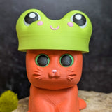 An orange cat figurine with green eyes wearing a bright green frog hat featuring cartoon eyes and a smiling face.