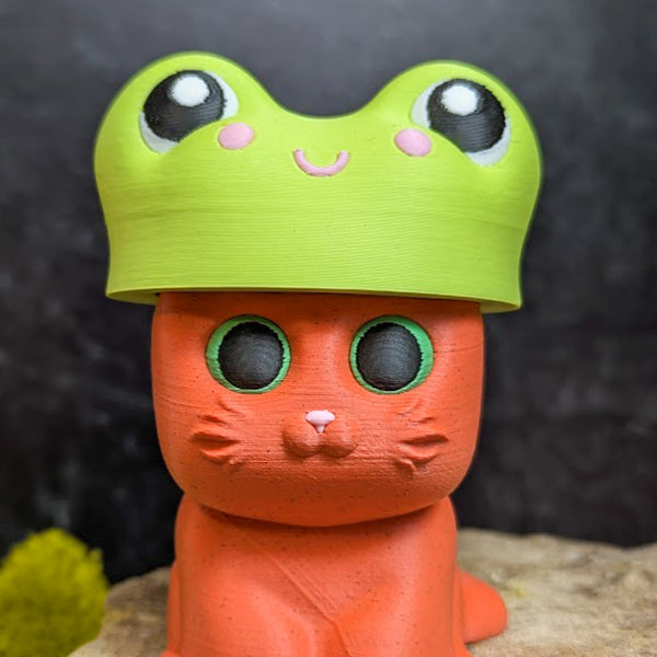 An orange cat figurine with green eyes wearing a bright green frog hat featuring cartoon eyes and a smiling face.