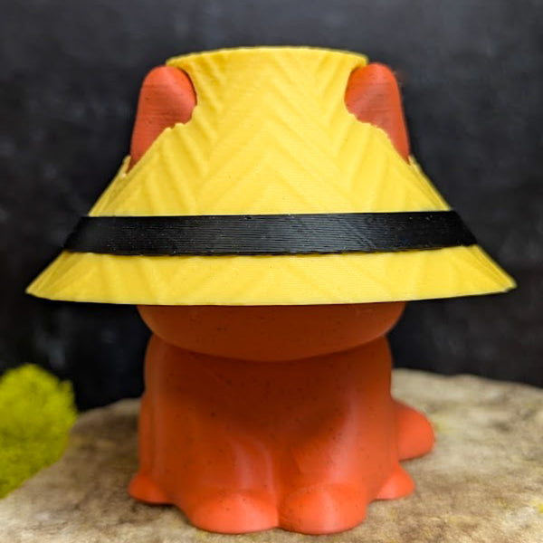An orange cat figurine mostly obscured by a wide yellow lampshade hat with a black band and cutouts for the ears.