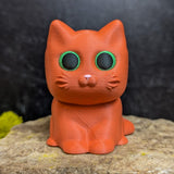 An orange cat figurine with wide green eyes and no hat, showing its small pointed ears and smooth round head.