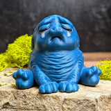 Crying blue Squonk figurine with wrinkled body, sitting on a stone with moss accents.
