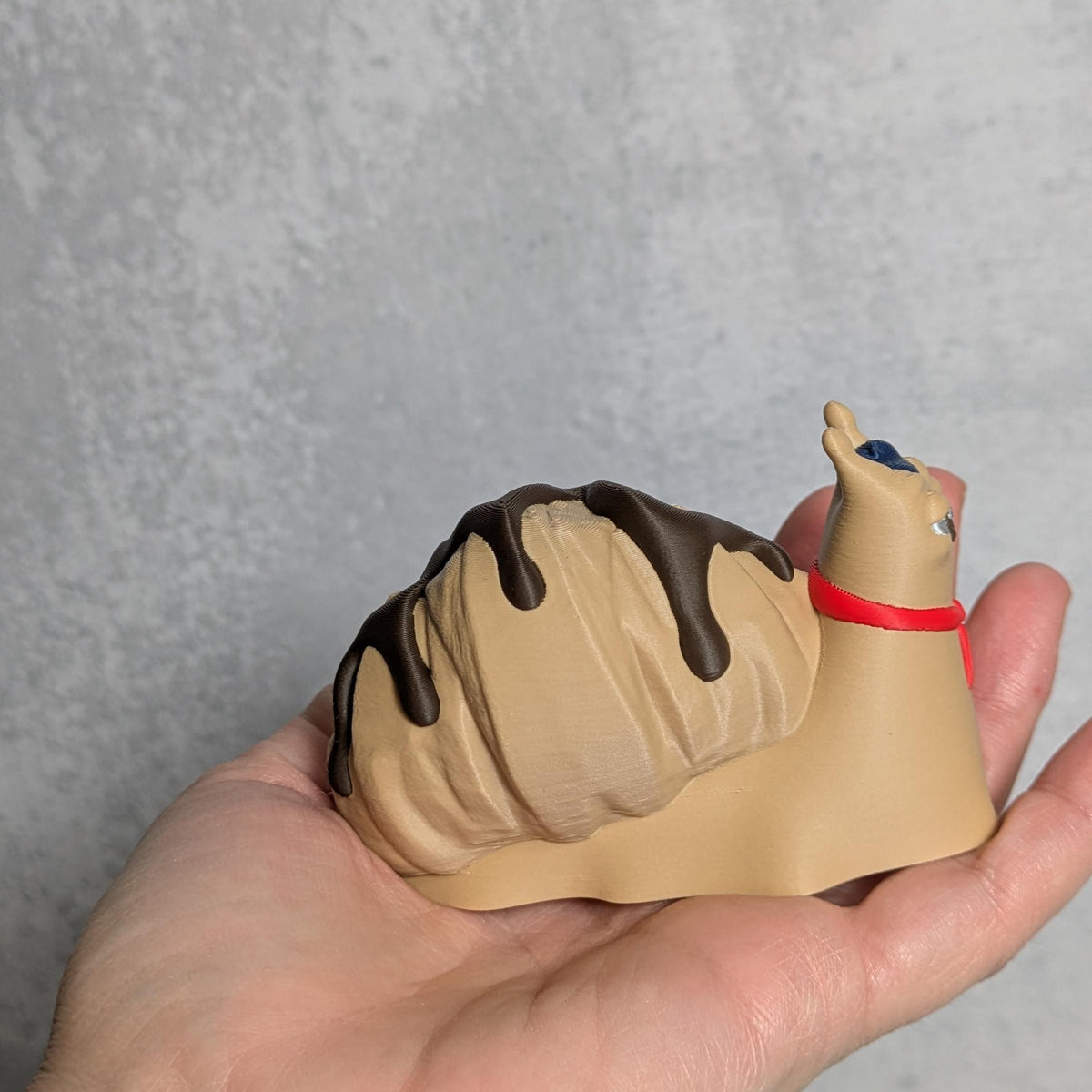 Angled view of Le Escargôutier™, a croissant snail figurine with a curled pastry shell and beady eyes, held in one hand.