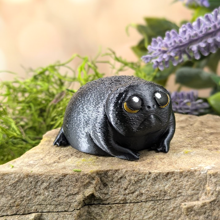 Angled view of a sad black rainfrog figurine with shiny eyes and a round body, sitting on a rock with greenery in the background.