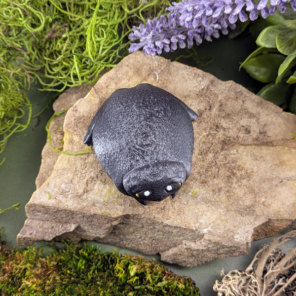 Overhead view of a frog figurine revealing its rounded shape, short limbs, and centered face, nestled on a mossy rock.
