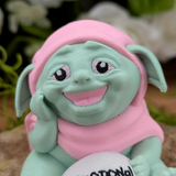 Close-up of a pastel green goblin figurine with a pink hood, smiling warmly with one hand resting on its cheek. Its large eyes have pink eyelids and a friendly expression.