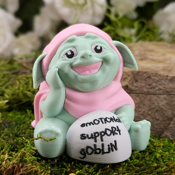 Front-facing view of a pastel green Emotional Support Goblin figurine wearing a pink hood, sitting on mossy ground next to a stone that says "emotional support goblin" in handwritten text.