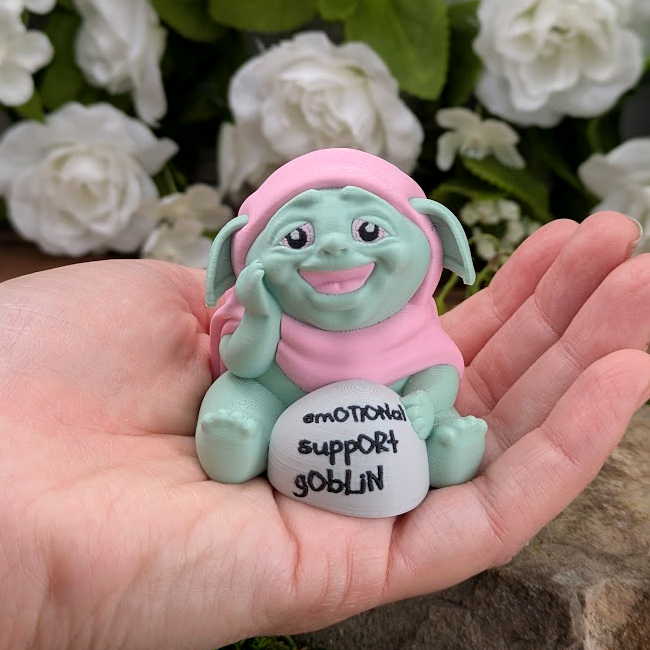 The Emotional Support Goblin figurine sitting in an open palm, showing its small scale. The goblin wears a pink hood and rests its hand on a labeled stone.