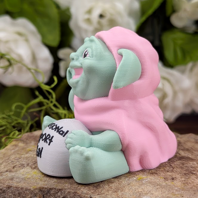 Side view of the goblin figurine from the left, showing its joyful face turned slightly upward. It’s perched on a stone surface with greenery in the background.