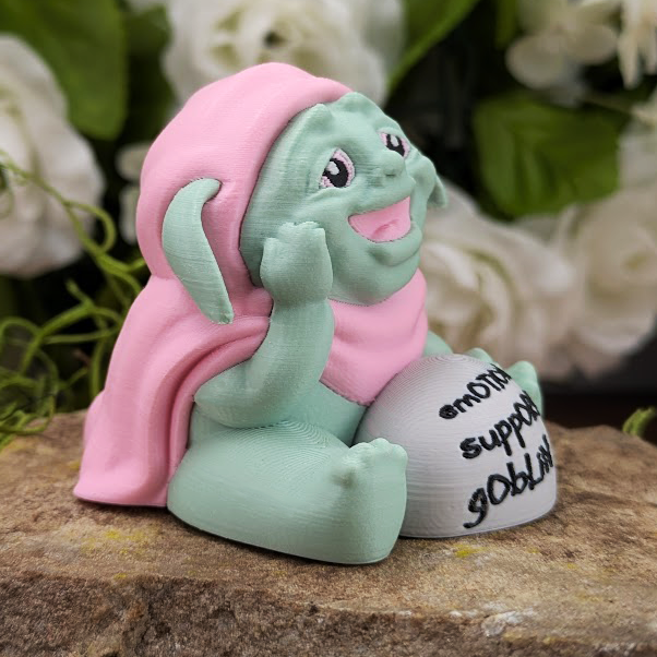 Angled right-side view of the Emotional Support Goblin, showing its cheerful face and relaxed pose on a mossy and stone background.